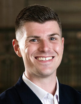 A white man with short dark hair wearing a white button-down shirt and a dark sports jacket.