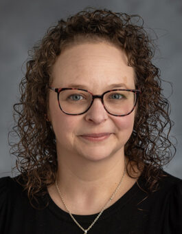 A white woman with dark curly shoulder length hair wearing a gold necklace and black blouse.