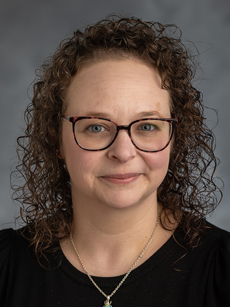 A white woman with dark curly shoulder length hair wearing a gold necklace and black blouse.