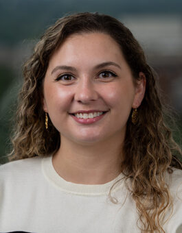 A white woman with long, curly blone hair wearing a white sweater and gold earrings.