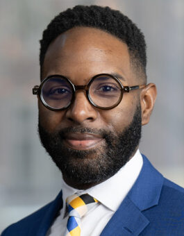 A black man with short dark hair and beard wearing with circular glasses, a brightly colored tie and a blue sports jacker