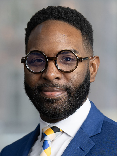 A black man with short dark hair and beard wearing with circular glasses, a brightly colored tie and a blue sports jacker