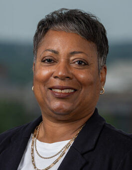 A black woman with short hair wearing gold earrings and necklaces and a dark blazer.
