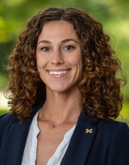 A white woman with brown curly hair wearing a dark blazer with a block M pin.