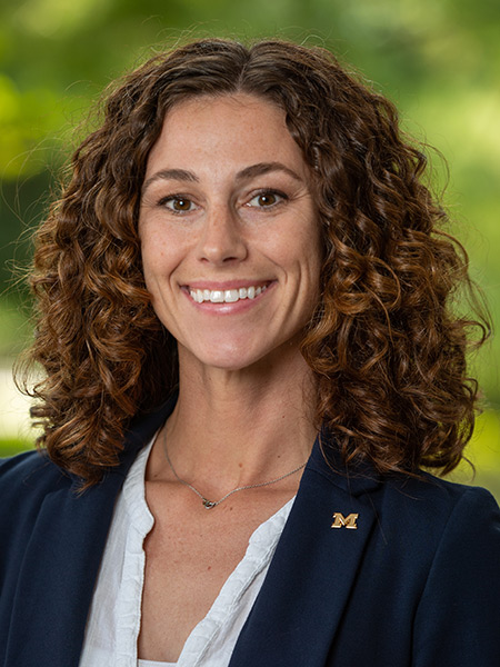 A white woman with brown curly hair wearing a dark blazer with a block M pin.