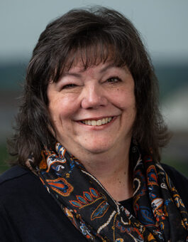 A woman with dark shoulder length hair wearing a patterned scarf and dark blouse.