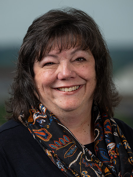 A woman with dark shoulder length hair wearing a patterned scarf and dark blouse.