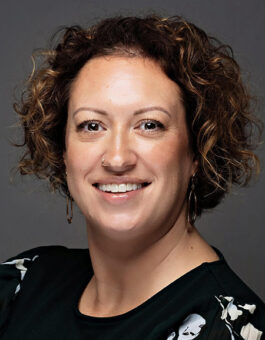 A woman with curly brown highlighted hair with a noise ring and earrings wearing a black floral blouse.