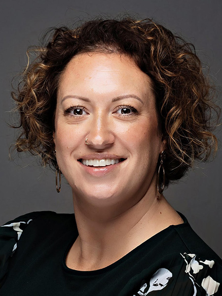 A woman with curly brown highlighted hair with a noise ring and earrings wearing a black floral blouse.