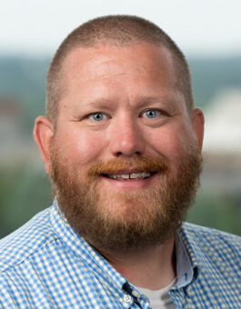 A white man with short hair and a red beard wearing a checked blue button-down.