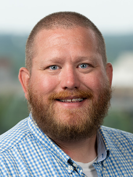 A white man with short hair and a red beard wearing a checked blue button-down.