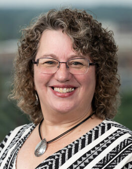 A white woman with shoulder length curly light brown hair and glasses waring an necklace and a black and white patterned shirt.