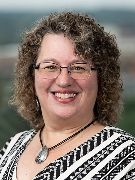 A white woman with shoulder length curly light brown hair and glasses waring an necklace and a black and white patterned shirt.
