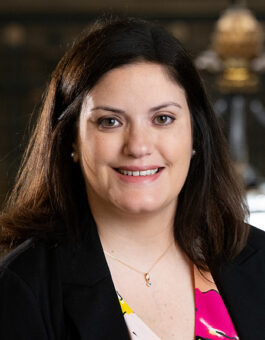 A woman with long dark hair wearing a brightly colored blouse and a dark blazer.