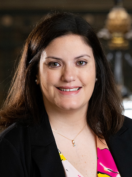 A woman with long dark hair wearing a brightly colored blouse and a dark blazer.