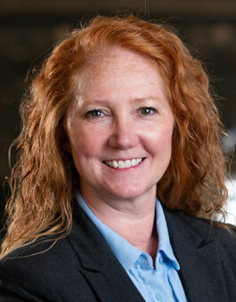 A white woman with long, red wavy hair wearing a blue button down and and dark blazer.