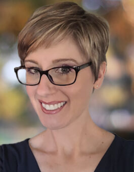 A white woman with short blonde hair wearing glasses and a dark shirt.