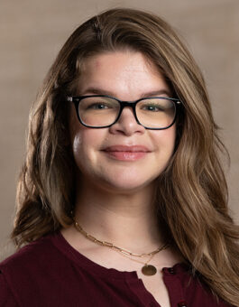 A white woman with long light brown hair and glasses wearing necklaces and a maroon blouse.