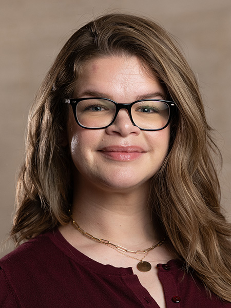 A white woman with long light brown hair and glasses wearing necklaces and a maroon blouse.