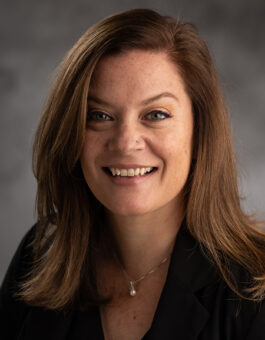 A white woman with long brown hair wearing a nose ring and pearl pendant and dark blouse.
