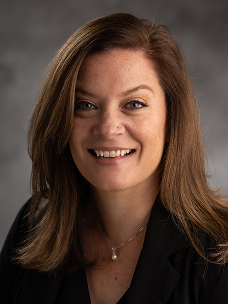 A white woman with long brown hair wearing a nose ring and pearl pendant and dark blouse.