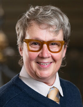 A person with gray short hair and orange plastic glasses wearing a dark sweater and gold tie.