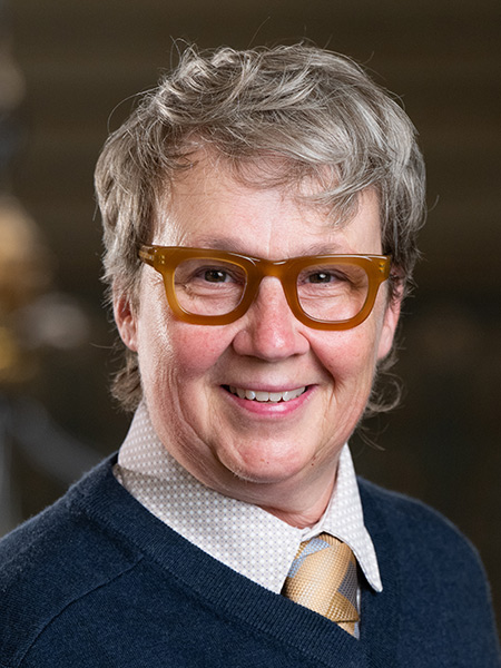 A person with gray short hair and orange plastic glasses wearing a dark sweater and gold tie.