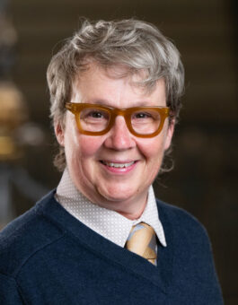 A person with short gray hair wearing brown glasses with a sweater and tie.