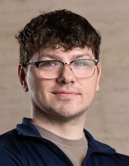 A white man with short curly dark hair wearing glasses and a sports zip up