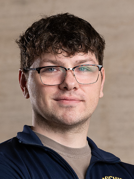 A white man with short curly dark hair wearing glasses and a sports zip up