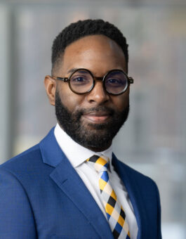 A black man with short dark hair, a trimmed beard and round glasses in a blue suit and multicolored tie.