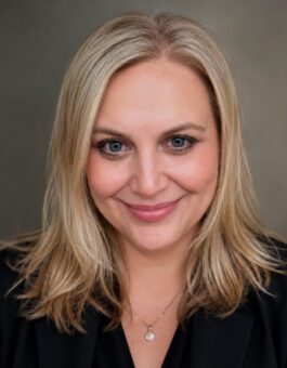 A white woman with blonde shoulder length hair wearing a pear pendant and a dark blouse.