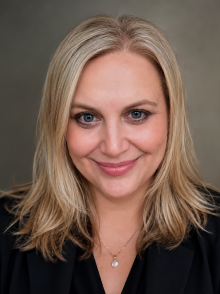 A white woman with blonde shoulder length hair wearing a pear pendant and a dark blouse.
