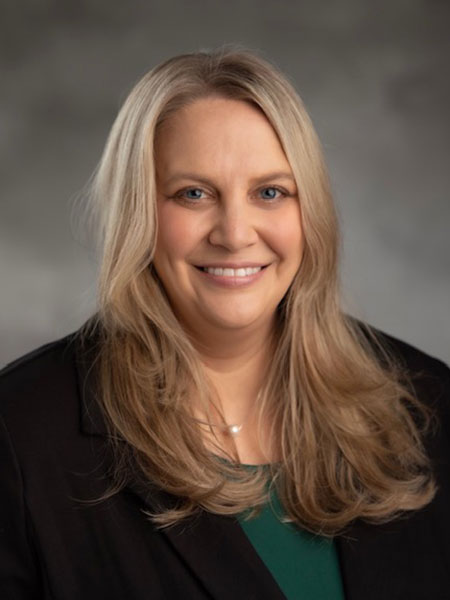 White woman with long blonde hair and wearing a dark shirt and suit and green shirt.