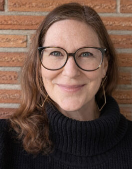 A white woman with long red hair and glasses wearing a black turtleneck sweater.