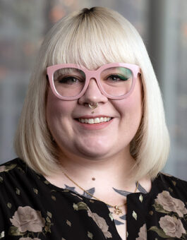 A white woman with blonde shoulder length hair wearing glasses, a nose ring and a dark floral blouse.