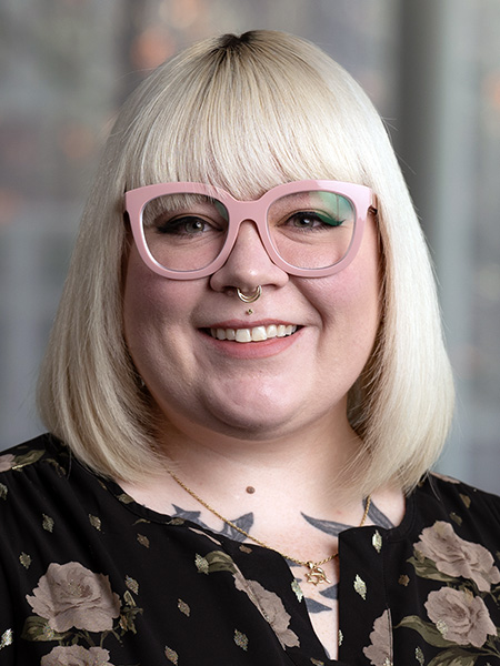 A white woman with blonde shoulder length hair wearing glasses, a nose ring and a dark floral blouse.