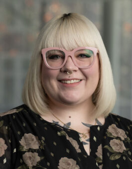 A white woman with a blonde bob and pink glasses in a dark floral dress.