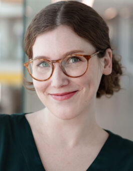 White woman with reddish brown wavy hair pulled back and brown eyeglasses in a dark green v-neck blouse.