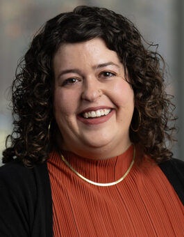 A white woman with curly, dark shoulder length hair wearing an orange blouse with a black cardigan and gold jewelry and a nose ring.