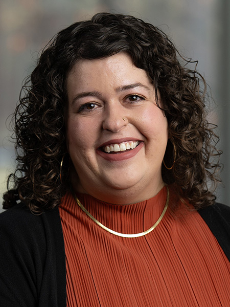 A white woman with curly, dark shoulder length hair wearing an orange blouse with a black cardigan and gold jewelry and a nose ring.