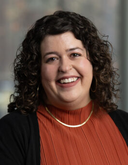 White woman with dark curly hair and a red blouse and black sweater.
