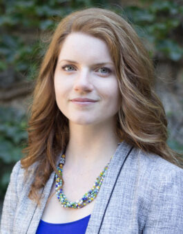 Jessica is a white woman with long, curly reddish brown hair with a white streak at the front and blue eyes. She wears a blue top, grey blazer, and colorful beaded necklace while smiling in front of an ivy-covered stone wall.