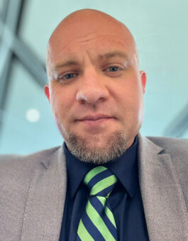 White man with a shaved head in a gray suit with a green striped tie.