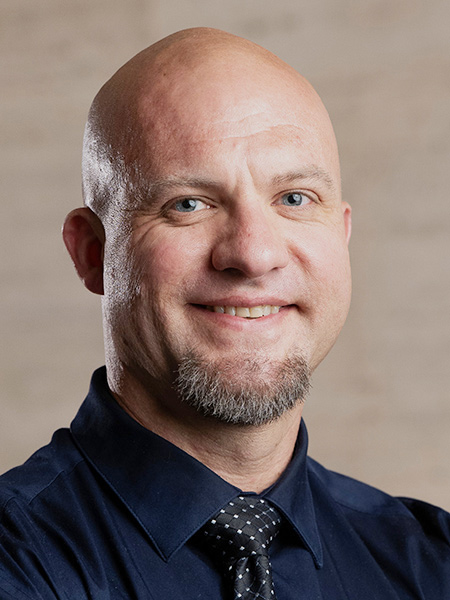 A white man with a shaved head and a goatee wearing a dark button-down and a polka dot tie.
