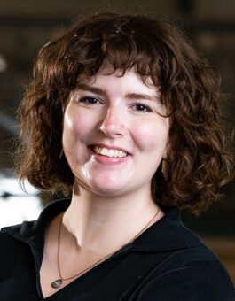 A white woman with a auburn, curly bob wearing a dark blouse and a necklace.