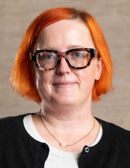 A white woman with bright orange hair wearing tortoise rimmed glasses and a dark cardigan.