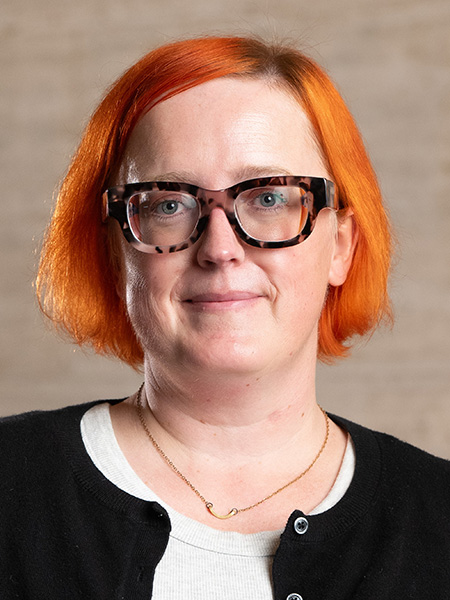A white woman with bright orange hair wearing tortoise rimmed glasses and a dark cardigan. 