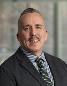 A hispanic man with short graying hair, a mustache, in a suit and tie.