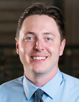 A white man with short dark hair wearing a baby blue button-down and tie